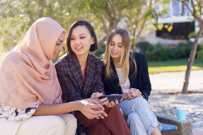 Smiling women sitting on outdoor park bench discussing something on smartphone