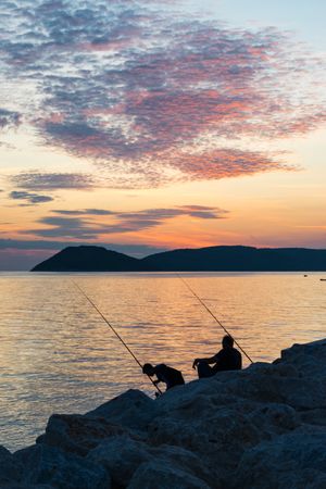 Silhouette of two fishermen sitting on rock near sea during sunset