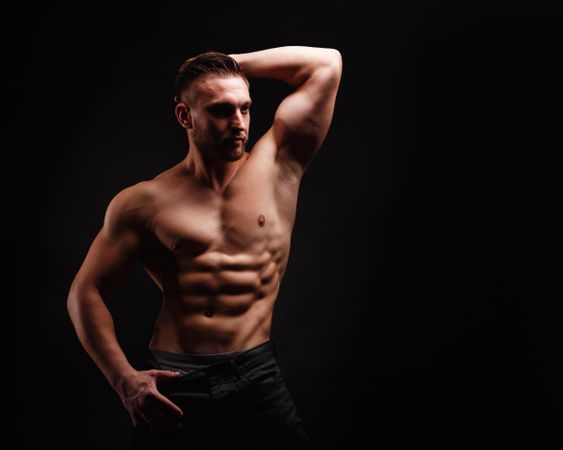 Bodybuilder practicing arm and abs poses in dark studio, copy space