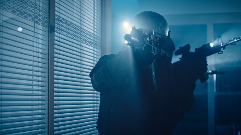 Close Up Portrait of Masked Armed SWAT Police Officer Storm a Dark Seized Office Building with Desks and Computers.