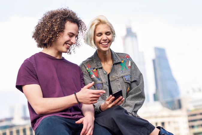 Smiling woman and happy curly haired man looking at phone while sitting on fence over river