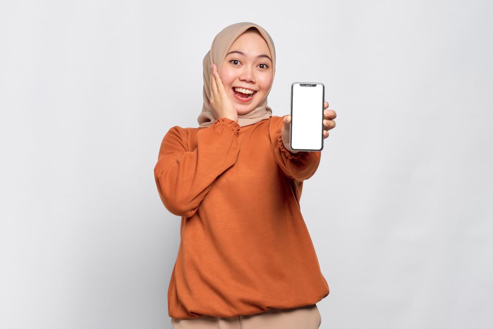 Surprised Muslim woman smiling holding up smartphone with mock up screen