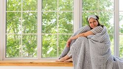 Woman sitting by window, enveloped in cozy gray blanket.