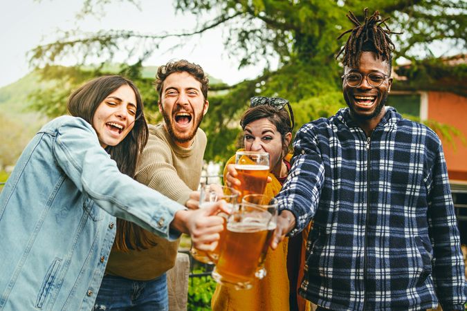 Cheers - multi-ethnic group of happy people toasting joining beer mugs and laughing looking at the camera