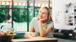 Portrait of Young Adult Woman with Blond Hair Wearing Gray V-Neck T-Shirt, Leaning on a Table and Smiling