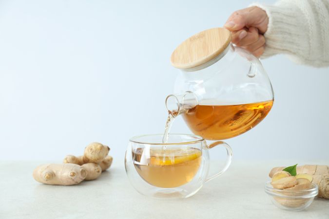Cup and teapot with tea in female hand, ginger on light background, space for text