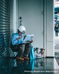Older man with helmet sitting on a sidewalk reading newspaper 0LJDD4