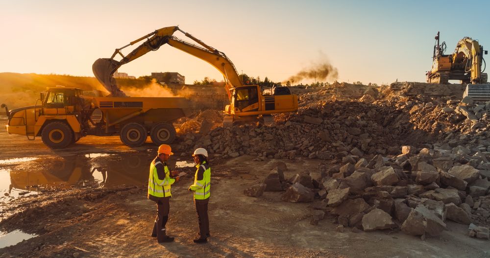 Aerial Drone Shot Of Construction Site On Sunny Evening: Male Civil Engineer Talking To Hispanic Female Urban Planner and Using Tablet. Industrial Excavator Loading Rocks Into A Truck.