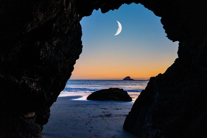 Crescent moon seen between rocky arch on quiet beach at dusk