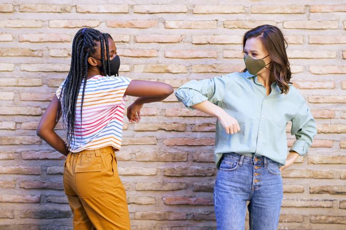Female friends greeting each other by tapping elbows in front of brick wall