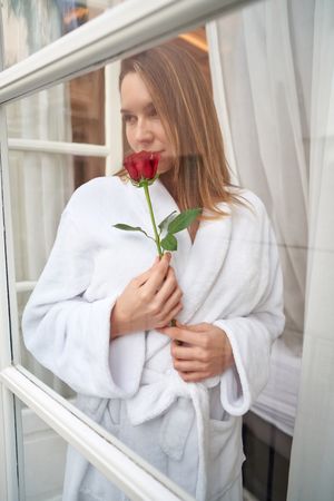 Young female in bathrobe standing at window with red rose