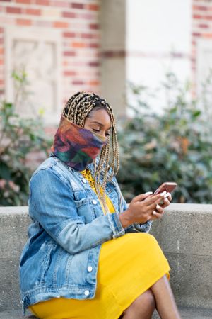 Woman with braids and mask sitting outside using her mobile phone