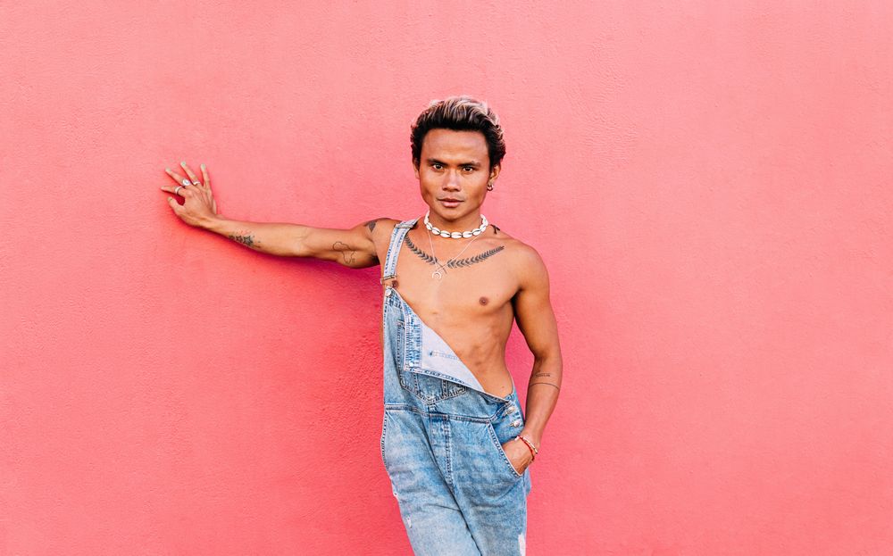 Male in open overalls looking at camera leaning against a pink wall