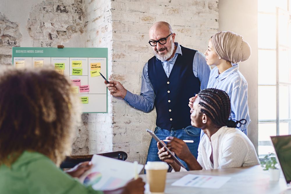 A mature bald man with a grey beard and glasses points at a canvas filled with sticky notes during presentation