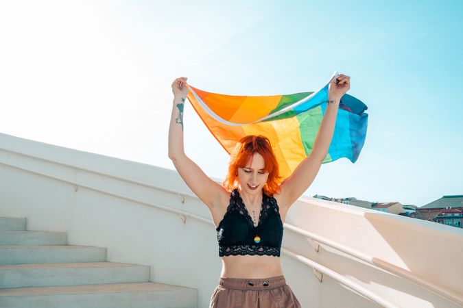 Young woman in dark lace bra waving rainbow flag under blue sky