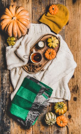 Flat-lay of beige knitted sweater, green woolen scarf, hat, decorative pumpkins, candle and tea