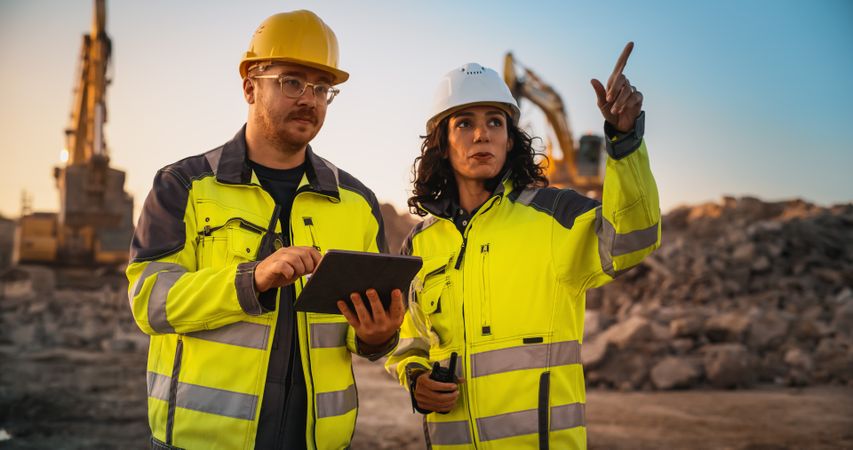 Male Civil Engineer Talking To Hispanic Female Inspector And Using Tablet Computer On Construction Site of New Building. Real Estate Developers Discussing Business, Excavators Working.