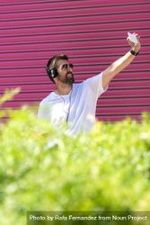 Latino man in headphones taking selfie with phone in front of pink wall 48w8Z5