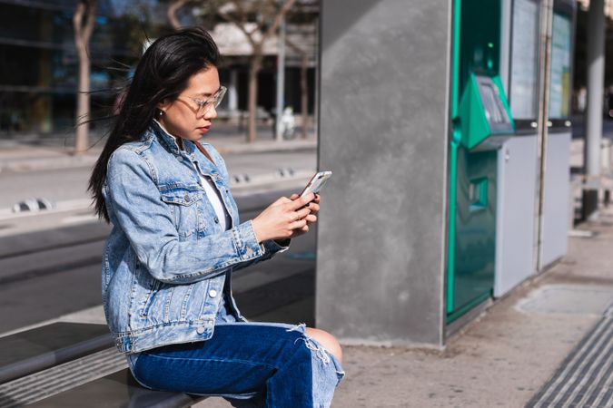Long hair brunette Asian woman using mobile phone while sitting on a bench in the street