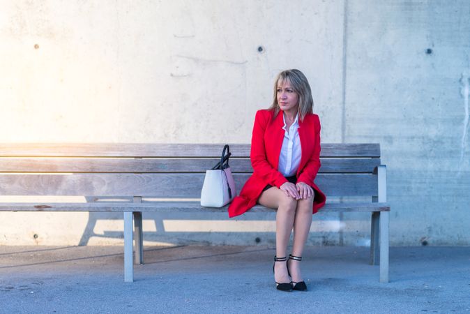 Blonde executive woman wearing red jacket sitting on a city bench while looking away with copy space