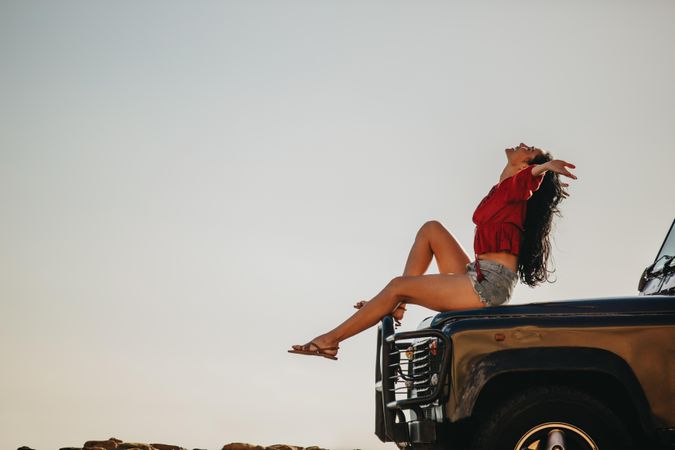 Young woman sitting on car hood with her hands outstretched and looking up