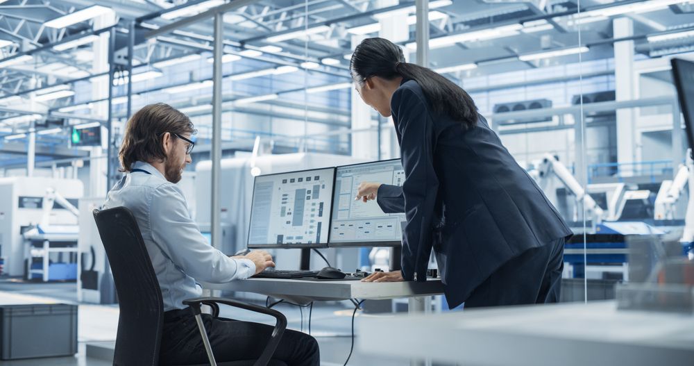 Handsome Engineer and a Beautiful International Business Manager Having a Conversation and Using Desktop Computer. Specialists Setting Up Production Values at an Electronics Assembly Plant