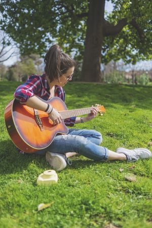 Front view of a young hipster woman wearing sunglasses, sitting on grass in a park while enjoying playing guitar
