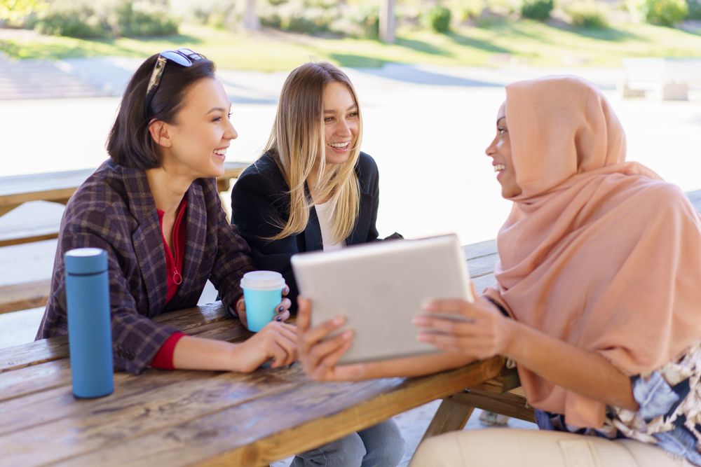 Three women on park bench with insulated mugs using digital tablet