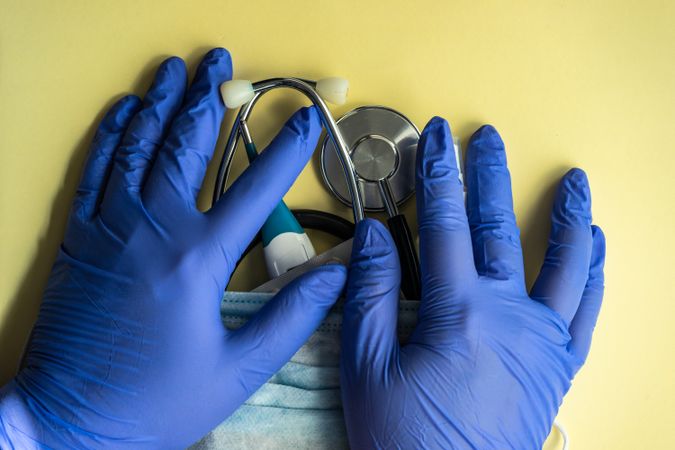 Top view of yellow table with stethoscope, facemask, protective gloves and digital thermometer