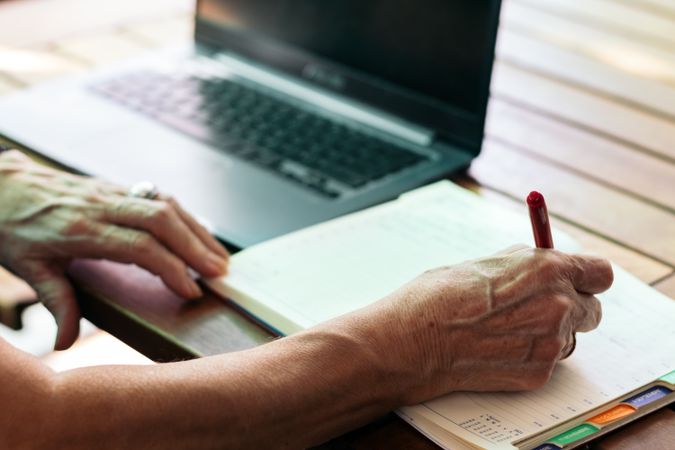 Unrecognizable older woman using a laptop and a notebook at home