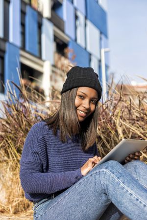 Female in hat and sweater sitting outside with tablet, vertical