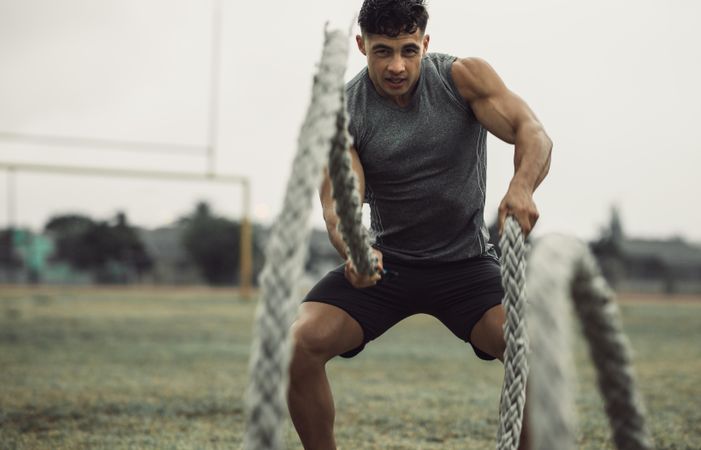 Fit young male athlete doing battle rope workout outdoors on a field