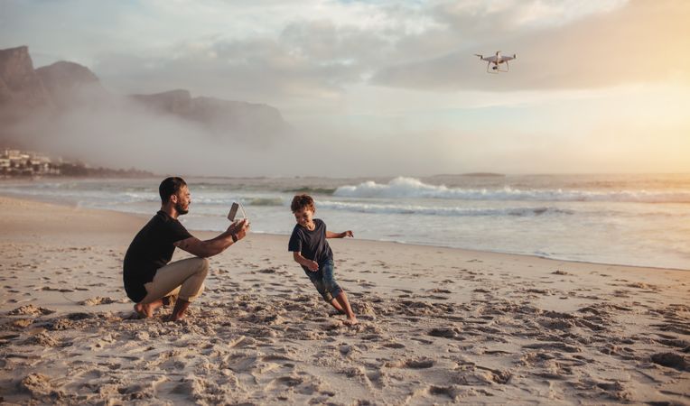 Handsome father flying drone and son running on beach