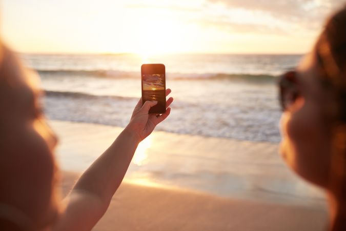 Two young women photographing sunset with smart phone on the beach