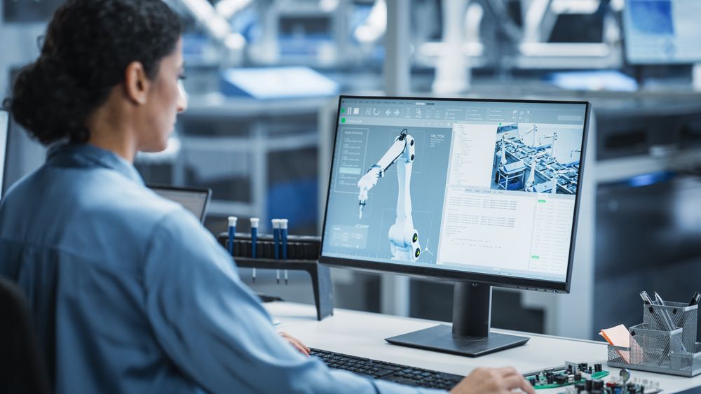 Hispanic Female Automation Technician Using Desktop Computer to Control Automated Production With Robotic Arms On Modern Electronics Factory.