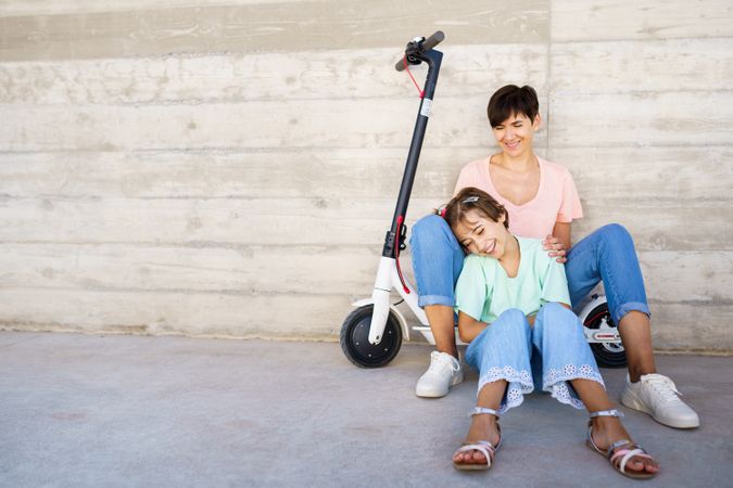Woman and preteen girl sitting on the ground outside posing with a scooter with copy space