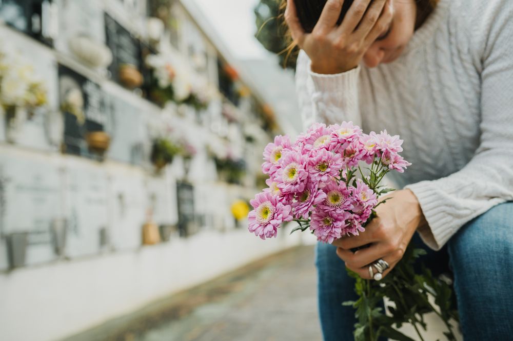 Grieving woman touching forehead while sitting near columbarium wall in cemetery