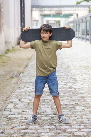 Young boy standing on the street while holding a skateboard behind his head while looking away