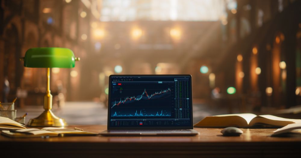Elegant Study Space With Green Desk Lamp and Open Books in Front of a Laptop Displaying Financial Charts