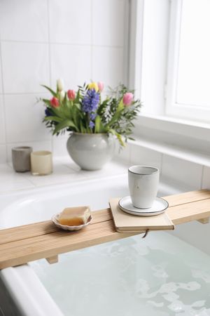 Cup of coffee, candles, book and soap on wooden board in elegant Scandinavian bathroom interior