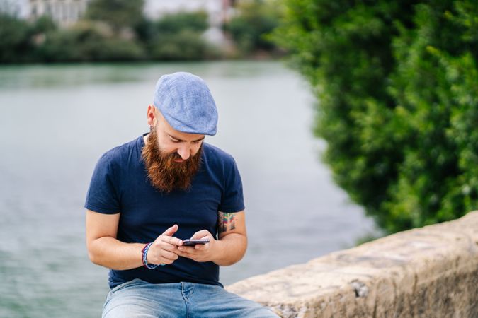 Bearded young man in casual wear using smartphone while walking on quay