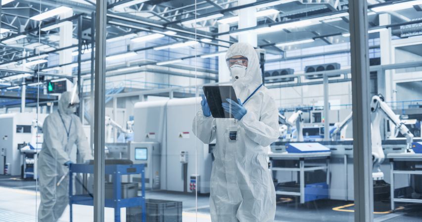 Portrait of a Female Worker Wearing Full-Body Protective Coveralls, Mask and Goggles at a Biotechnology Manufacturing Plant. Young Specialist Using a Tablet Computer, Overlooking Nanotech Production