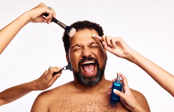 Hands of women shaving, applying makeup and cologne to excited bearded person