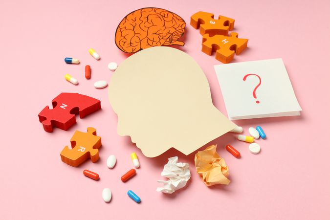 Flat lay paper cut out of side view of head with medications and puzzle pieces on pink background