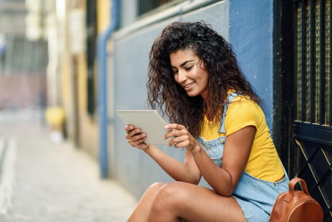 Smiling Arab woman looking at her digital tablet sitting outside a door, copy space