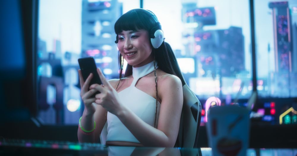 Smiling Asian Female Using Smartphone while Sitting Behind a Desk in a Futuristic Cyberpunk Office Space.