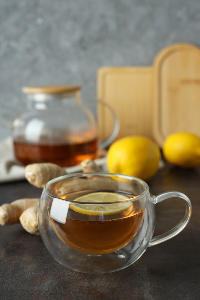Cup and teapot with tea, lemon and ginger on gray background