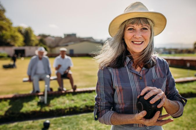 Mature woman in hat standing in a lawn holding a boules with friends in background
