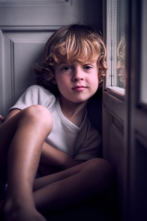 Tranquil little kid sitting near window and looking at camera