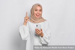 Asian Muslim woman in a bright studio shoot holding cell phone and pointing upwards 486wq0